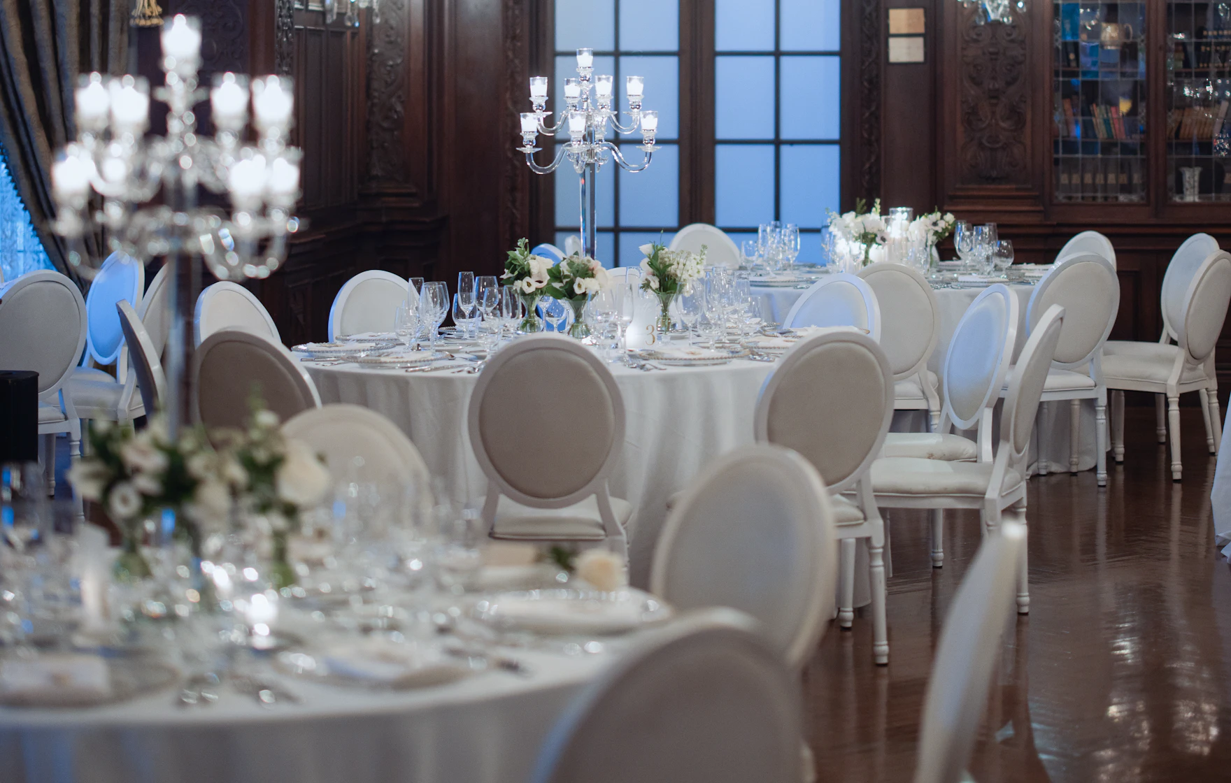 Zaal met gedekte ronde tafels, witte stoelen en kroonluchters, klaar voor de gasten